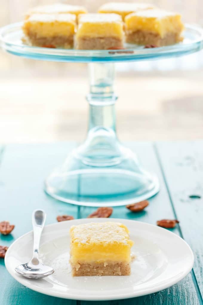 Pecan and Lemon Bars  on white plate with spoon on blue table with glass tray with lemon bars and pecans spilled on table#dessert