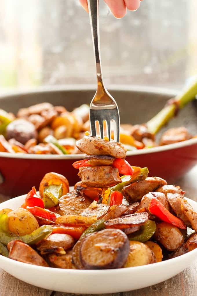 Creamer potatoes with sausage and pepers on white plate with fork on wooden table with pan full of same dish in the background