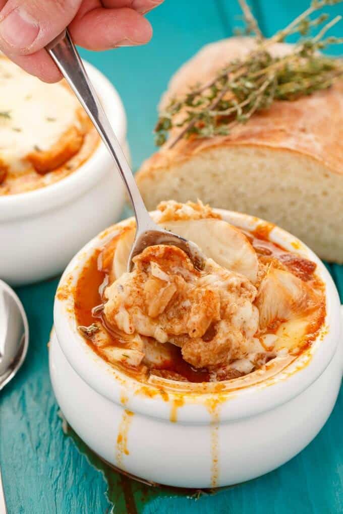 Massaman Curry French Onion Soup in white bowl, spoon held by hand  on blue table with loaf of bread and herbs