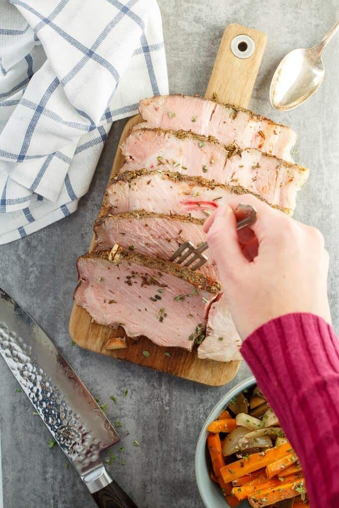 Pork Shoulder with Vegetables in the Slow Cooker on wooden pad picked on fork held by hand. Knife, spoon, cloth wipe and bowl of vegetable on gray table