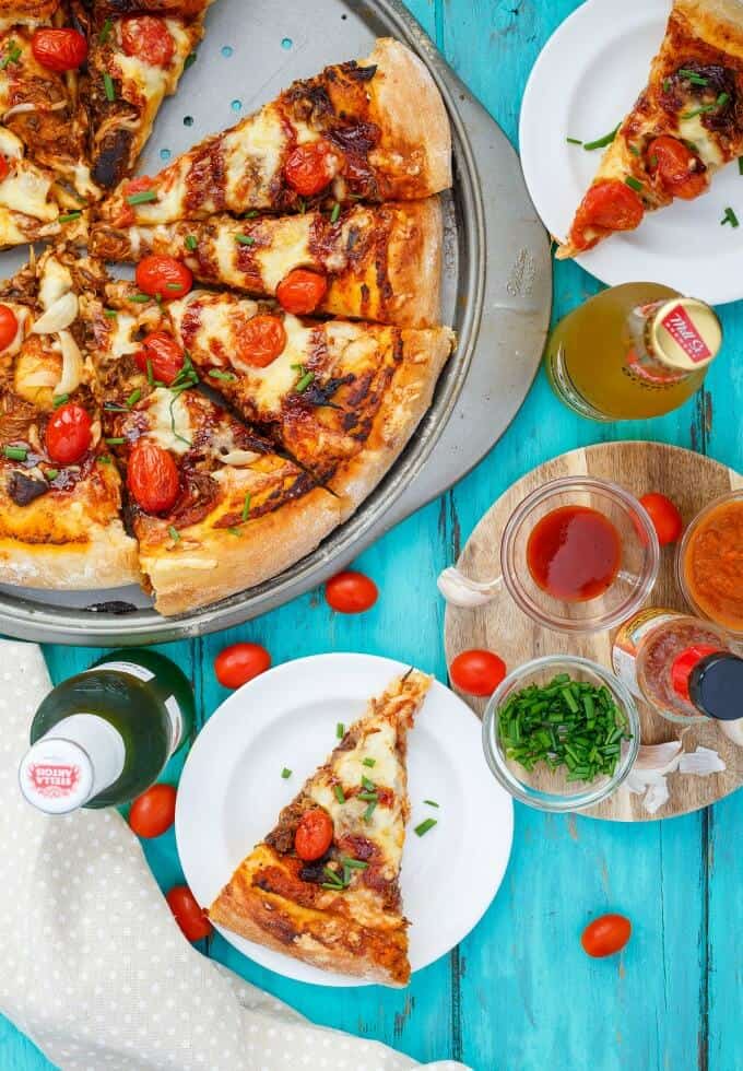 BBQ Pulled Pork Pizza using Leftovers on try, slice of pizza on white plate on blue table with beer bottle, sauces and veggies