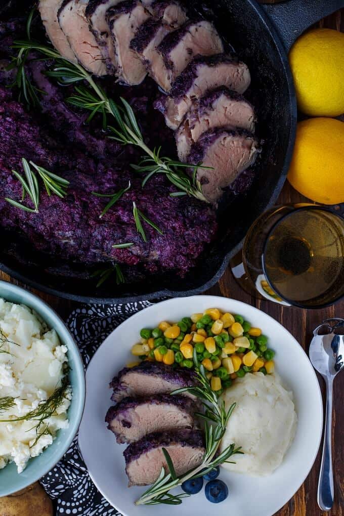 Blueberry Molasses Pork Tenderloin in baking pot and white plate, with side dish with veggies, frui, herbs, lemons, spoon, glass of wine, bowl of side dish