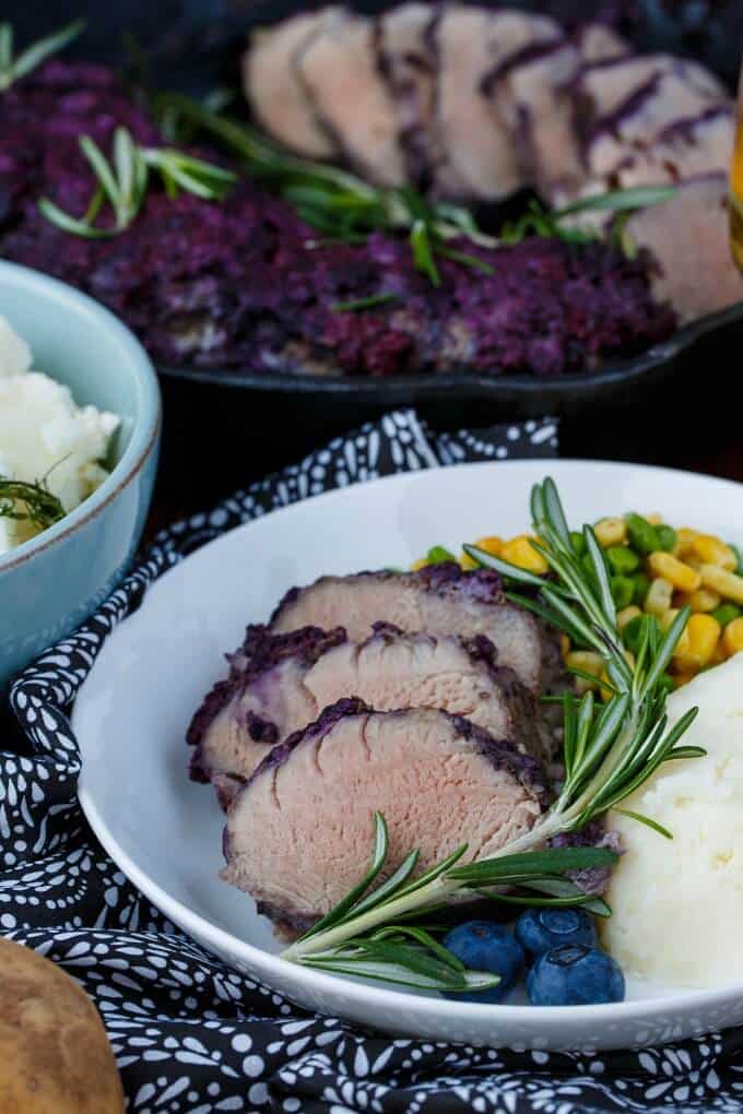 Blueberry Molasses Pork Tenderloin on white plate and baking pot with side dish, herd, veggies, fruits