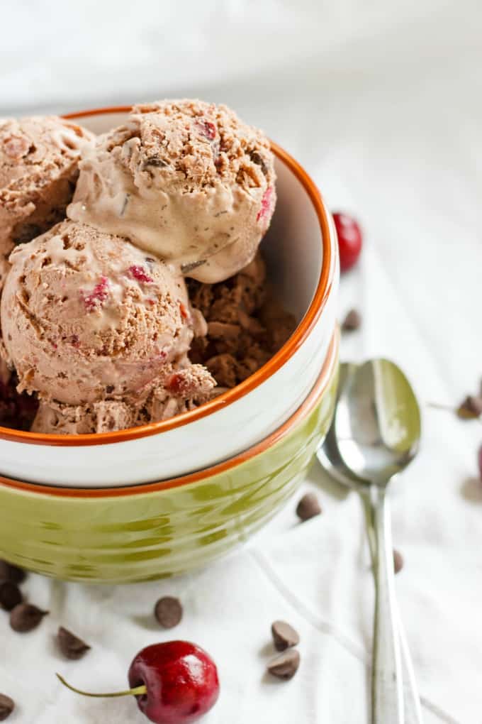 Cherry-Chocolate Ice Cream in pile of bowls on white table with spoons, chocolate chips and cherries