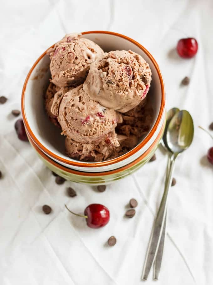 Cherry-Chocolate Ice Cream in pile of bowls on white table with spoons, chocolate chips and cherries