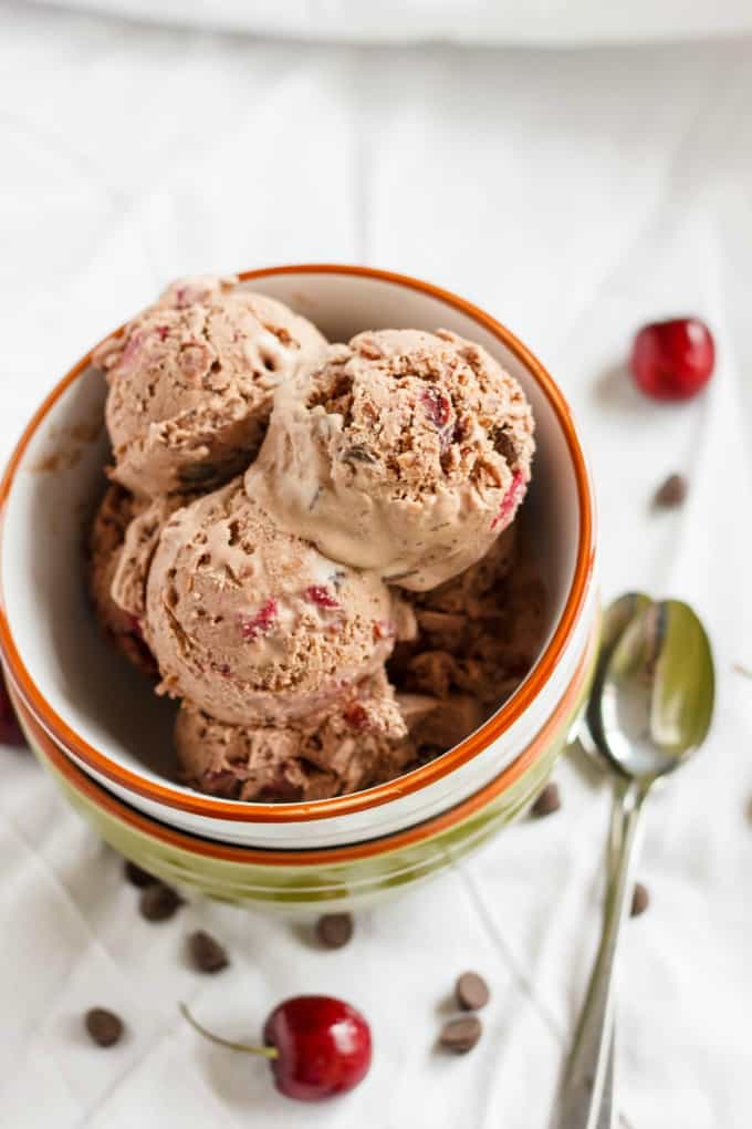 Cherry-Chocolate Ice Cream in pile of bowls on white table with spoons, chocolate chips and cherries