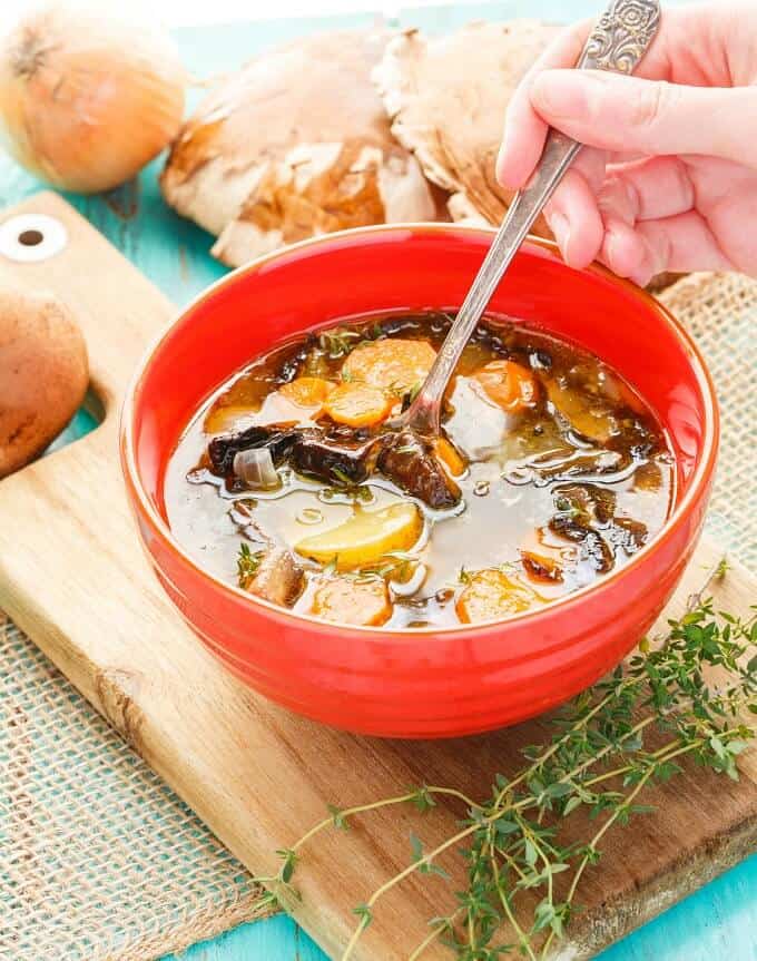 Portobello Mushroom Soup with Homemade Chicken Stock mixed by spoon held by hand on wooden pad next to herb, onions and mushrooms