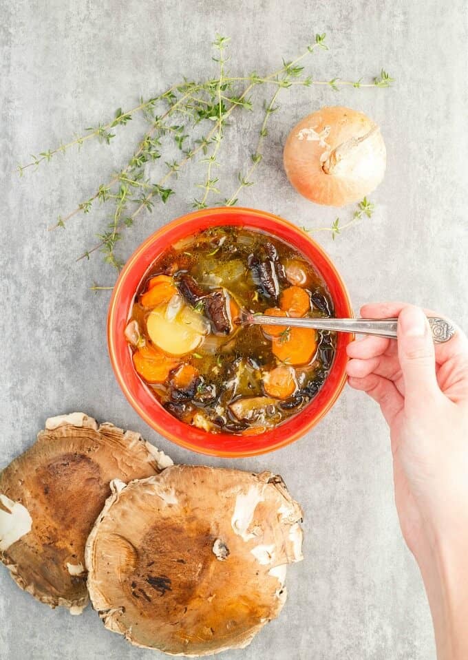 Portobello Mushroom Soup with Homemade Chicken Stock in red bowl mixed by spoon held by hand next tp herb, onion and mushrooms