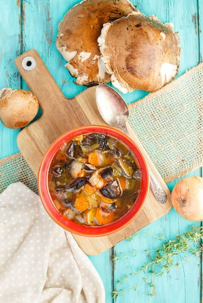 Portobello Mushroom Soup with Homemade Chicken Stock in red bowl on wooden pad next to spoon, mushrooms and onions