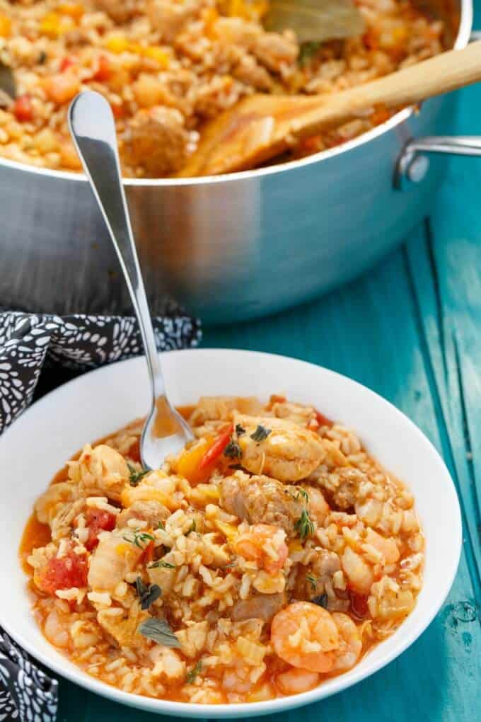 Jambalaya dish on white plate with spoon on blue table, jambalaya in pan with wooden spatula in the background