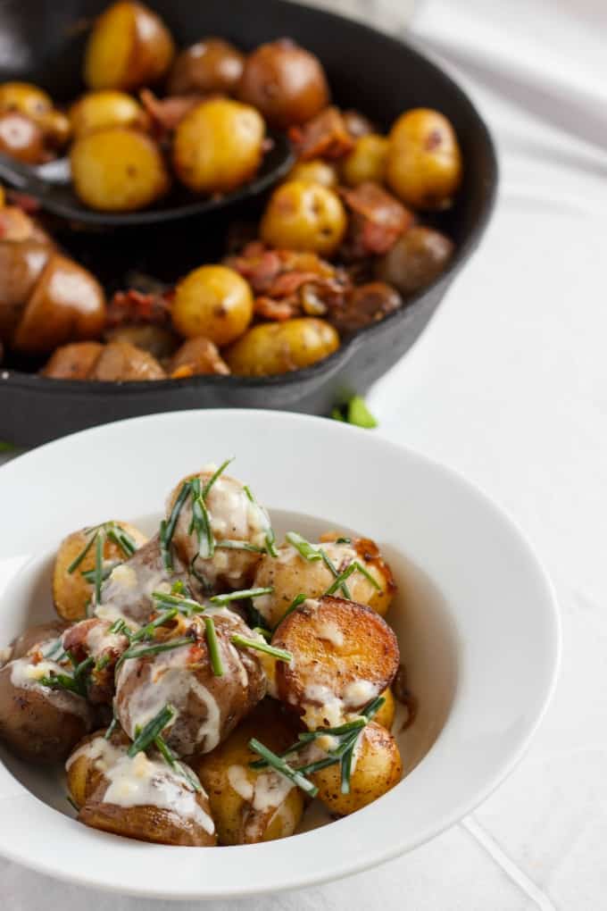 Pan-Fried Creamer Potatoes and Bacon with a Yogurt-Chive Sauce  on white plate on white table next to black pan full of same dish