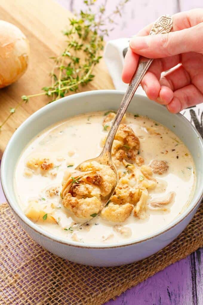 Roasted Cauliflower Chicken Soup in white bowl with spoon held by hand on purple table with wooden pad with onion and herbs