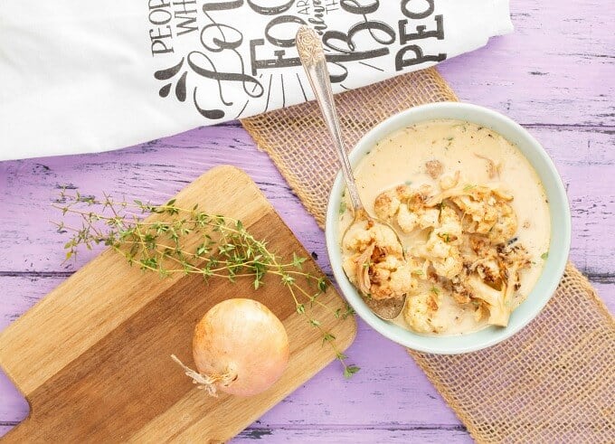 Roasted Cauliflower Chicken Soup in white bowl with spoon on purple table with wooden pad , onion, herbs and cloth wipe