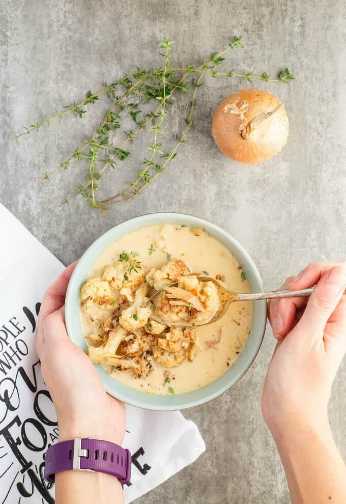 Roasted Cauliflower Chicken Soup in white bowl, hands holding spoon and bowl, on table with onion, herbs and cloth wipe