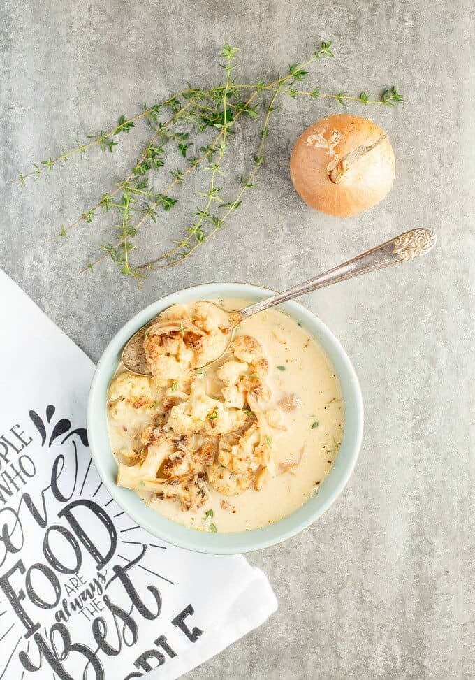 Roasted Cauliflower Chicken Soup in white bowl with spoon on table with onion, herbs and cloth wipe