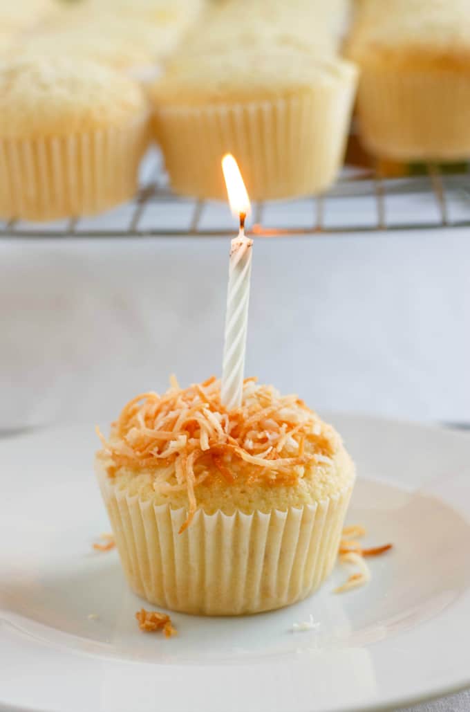 Toasted Coconut Cupcakes with Coconut Glaze with candle on white plate, cupcaked on baking grid in the background