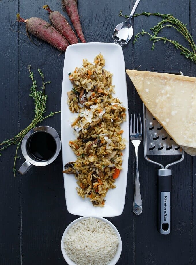 Carrot, Leek, and Mushroom Risotto on white tray on gray table with jar of sauce, fork, spoon, grater, cheese, carrots, herbs, bowl of rice