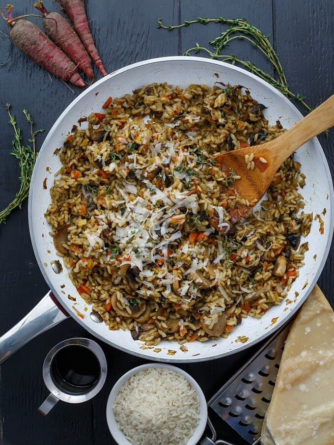 Carrot, Leek, and Mushroom Risotto on pan with wooden spatula on gray table with bowl of rice, jar of sauce, grater, cheese, herbs, carrots