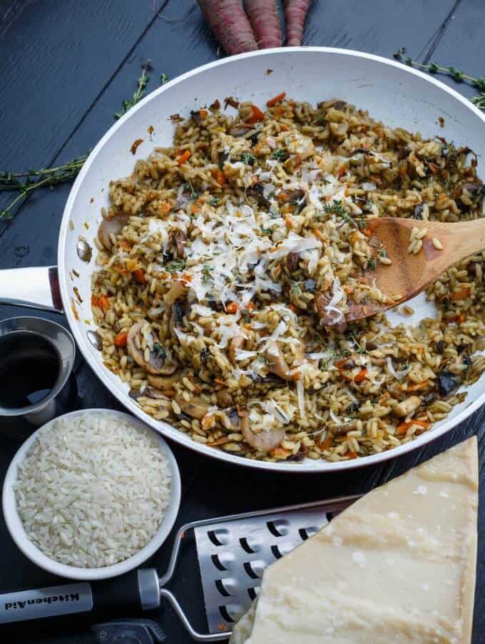 Carrot, Leek, and Mushroom Risotto on pan with wooden spatula on gray table with rice, grater, cheese, herbs, carrots, jar with sauce
