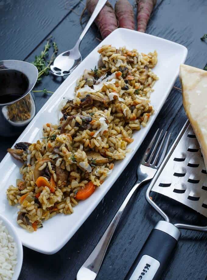 Carrot, Leek, and Mushroom Risotto on white tray on gray table with spoon, carrots, fork, grater, cheese, herb and jar with sauce