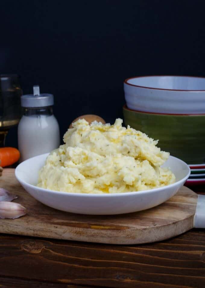 Herb and Garlic Cream Cheese Mashed Potatoes on white plate on wooden pad with garlic, bowls and jar of milk