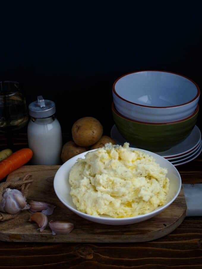 Herb and Garlic Cream Cheese Mashed Potatoes pon white plate on wooden pad with garlic, carrot, potatoes, bowls, plates and jar of milk