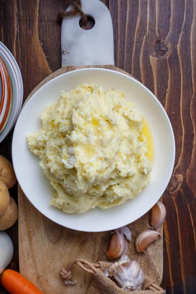 Herb and Garlic Cream Cheese Mashed Potatoes on white plate on wooden pad with garlic, carrot