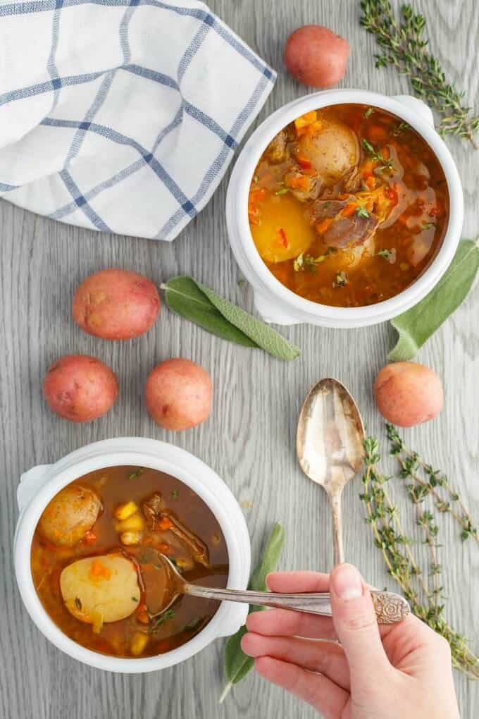 Homemade Turkey Soup with Creamer Potatoes in white bowl with spoons, potatoes, herbs, cloth wipe and hand holding spoon