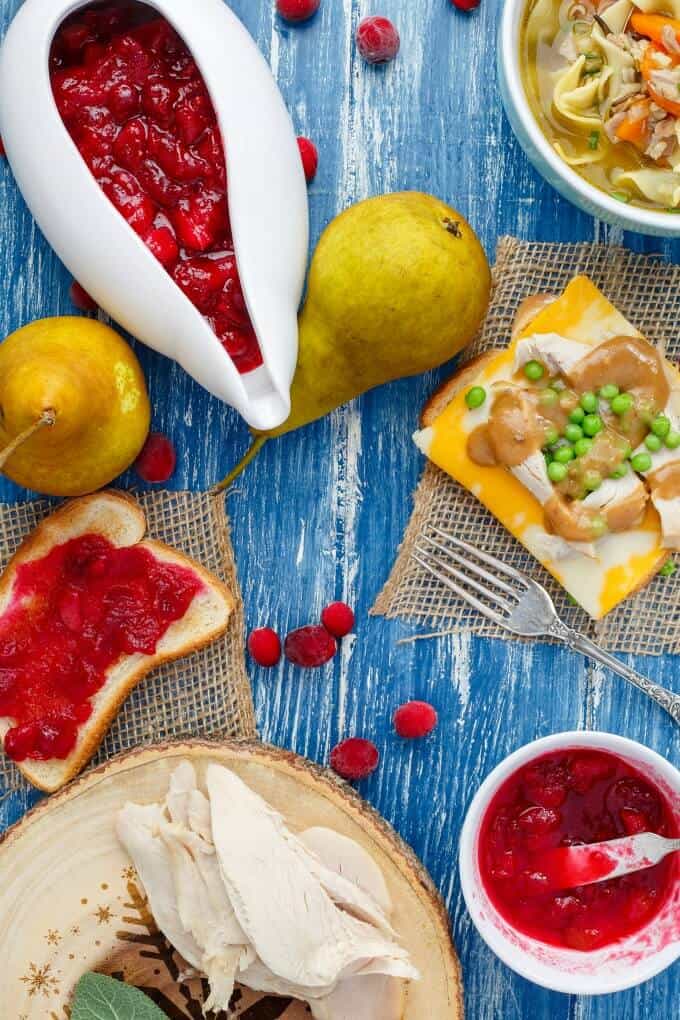 Pear Cranberry Sauce in white bowls with sandwiches, forks, peaches. wooden pad with turkey meat, bowl of dish, scattered cranberries on blue table