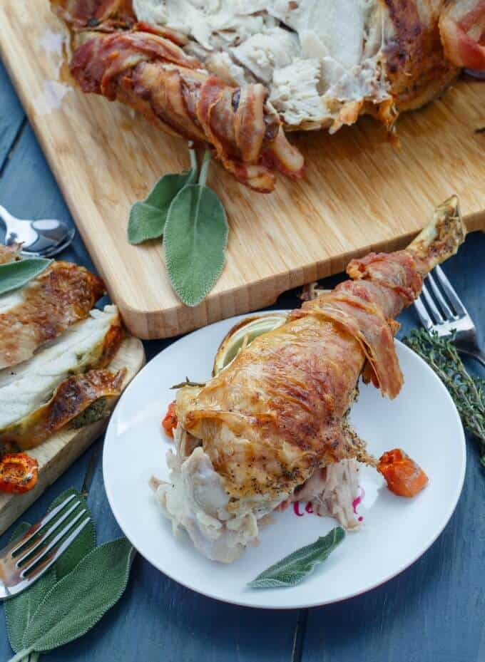 Bacon-Wrapped Whole Turkey with Herb Butter on white plate and wooden pad with forks and herbs