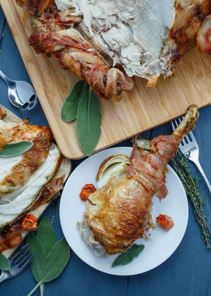 Bacon-Wrapped Whole Turkey with Herb Butter on white pad and white plate on blue table with spoon, forks and veggies