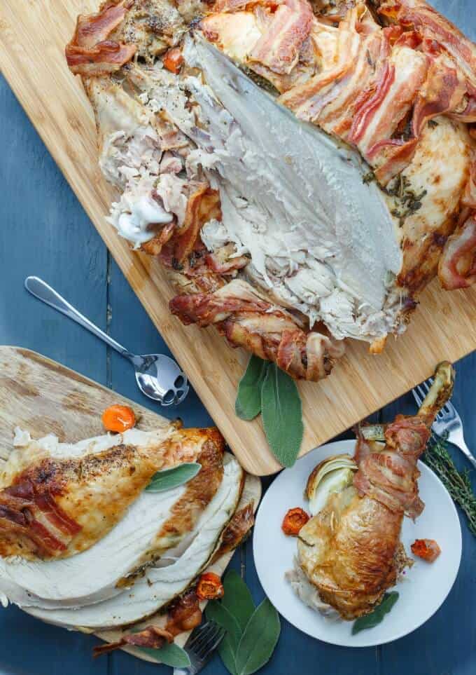 Bacon-Wrapped Whole Turkey with Herb Butter on wooden pads and white plate next to spoon, fork , herbs on gray table