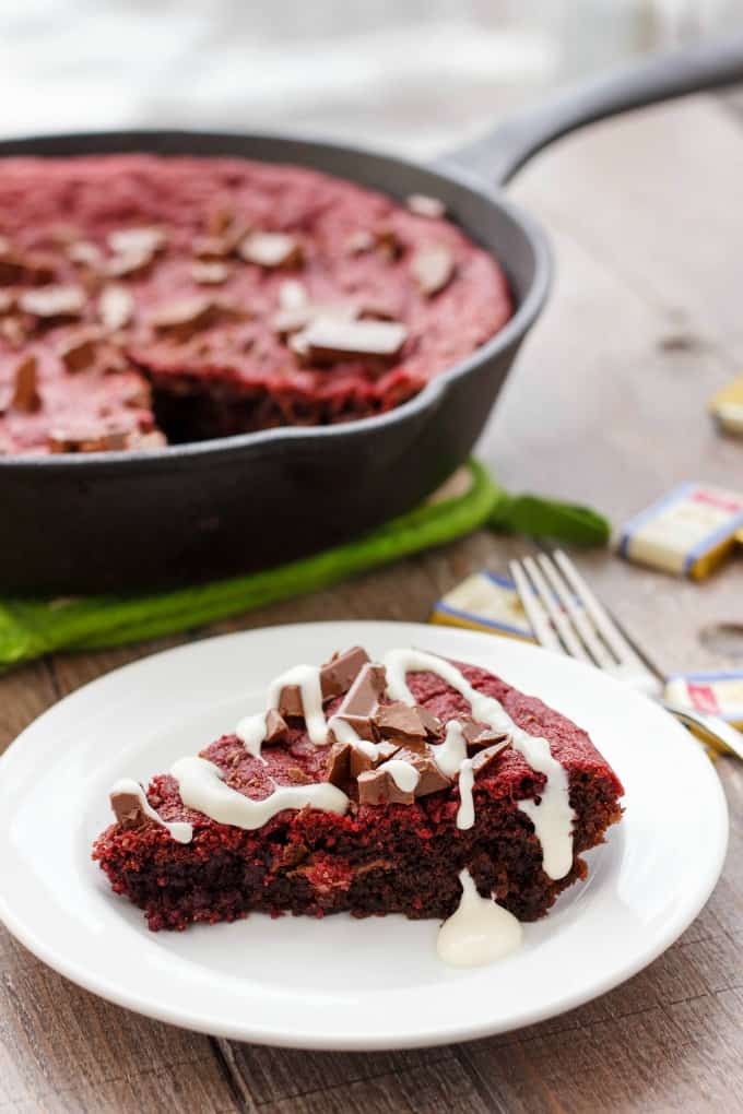 Cast iron red velvet cookie skillet in black pan, slice of cookie on white plate next to fork on wooden table