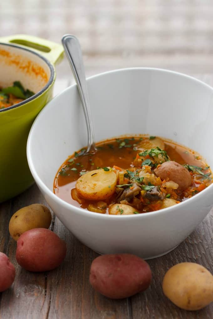 Simple Vegetable Soup with Creamer Potatoes in white bowl with spoon on wooden table with spilled potatoes and green pot