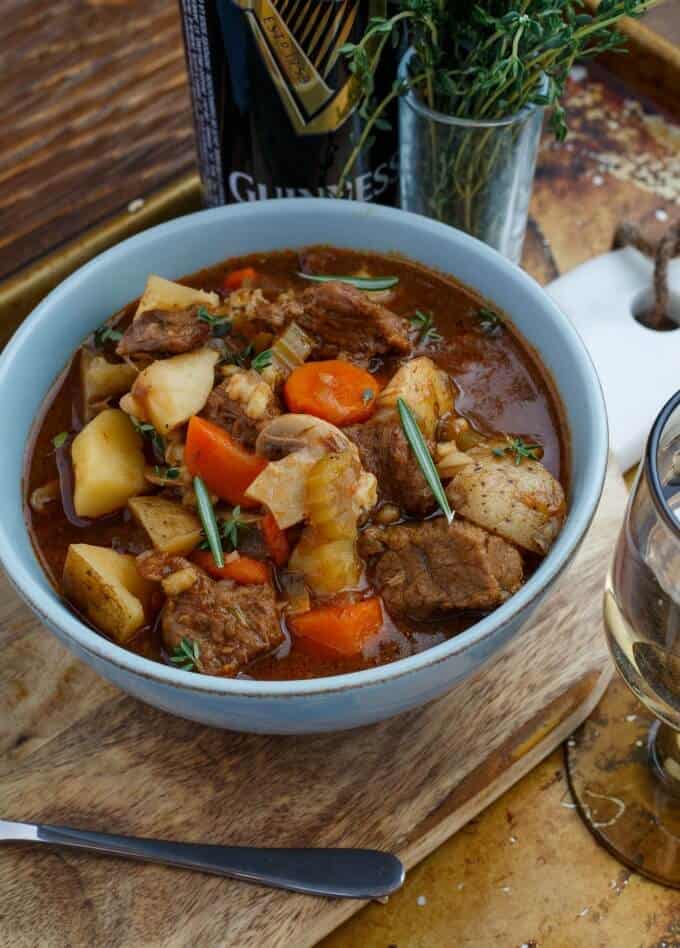 Beef Stew with Barley in blue bowl on wooden pad with spoon, herbsm glass and bottle