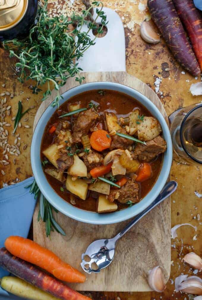 Beef Stew with Barley in blue bowl on wooden pad with spoon, carrots, herbs, glass and beer