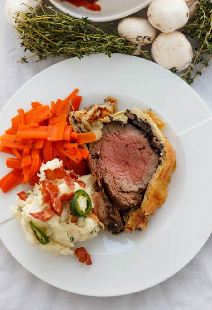 Beef Wellington with Bacon and Button Mushrooms , pepper, veggies on white plate next to mushrooms and herbs#beef