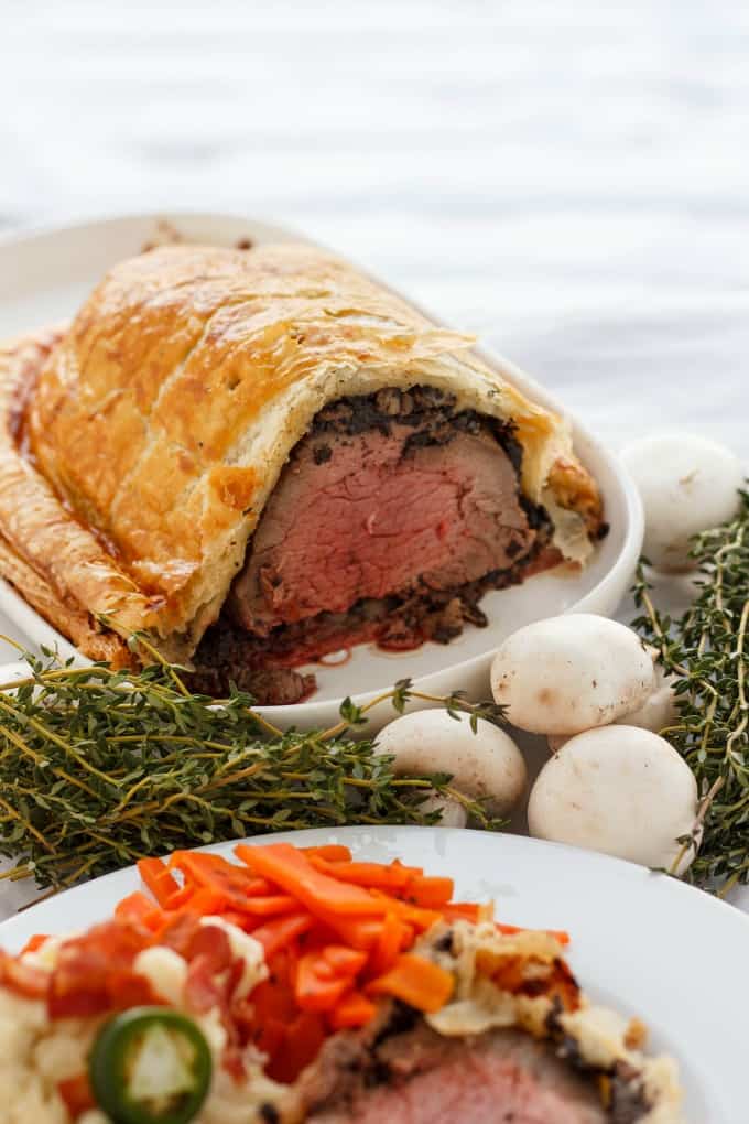 Beef Wellington with Bacon and Button Mushrooms on white tray and palte with veggies, peppers next to mushrrom and herbs #beefwellington