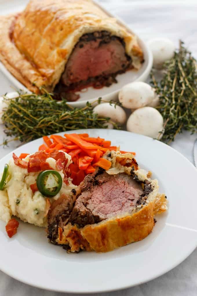 Beef wellington with bacon and buttom mushrooms on white plate, rest of dish,mushroom and herbs in the background