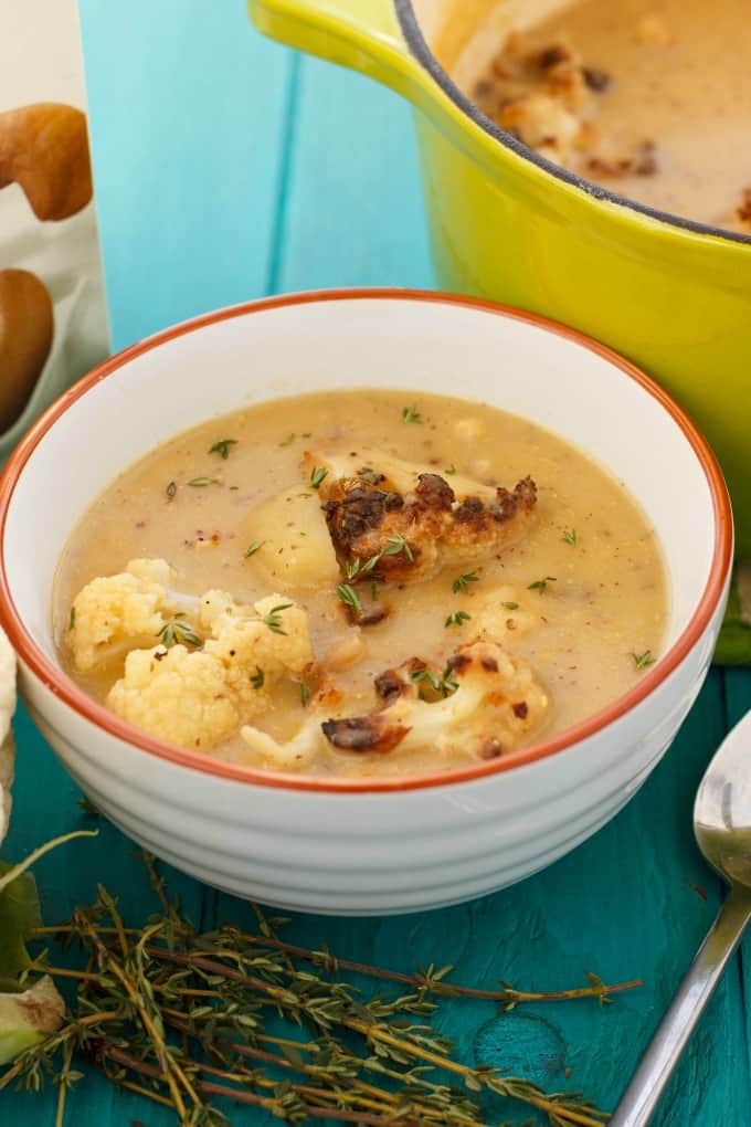 Creamy Vegan Cauliflower Soup with Cashew Milk in white bowl on blue table with herb, spoon and yellow pot#healthy