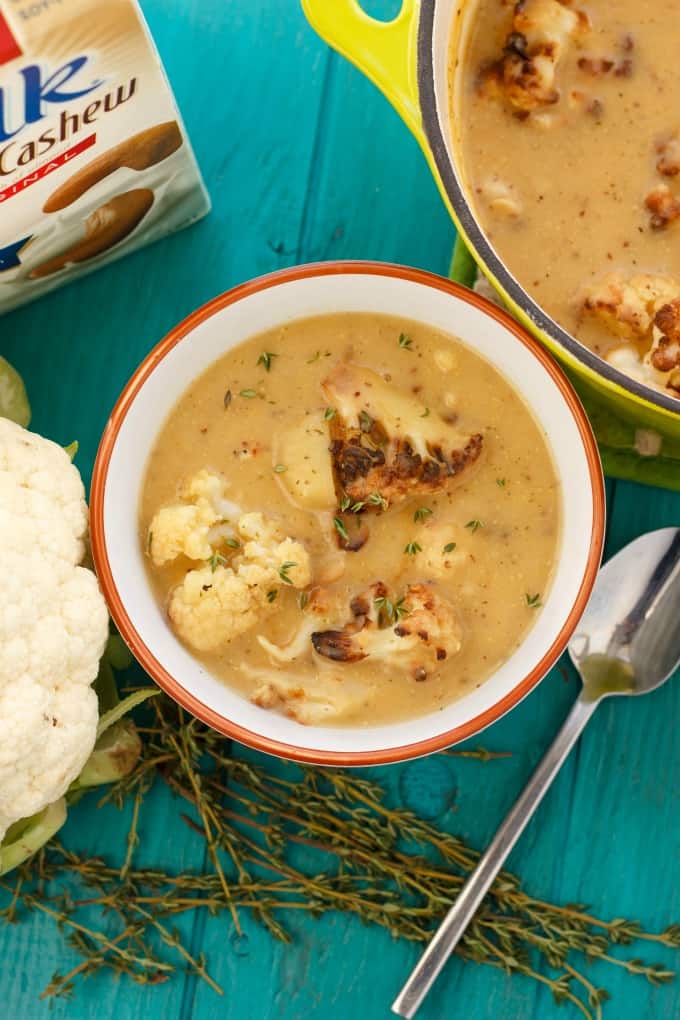 Creamy Vegan Cauliflower Soup with Cashew Milk in white bowl on blue table with spoon, herb, cauliflower, pot and cashew milk#vegan