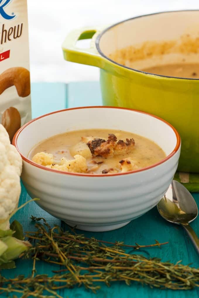 Creamy Vegan Cauliflower Soup with Cashew Milk in white bowl on blue table with spoon, herb, cauliflower, green pot and cashew milk