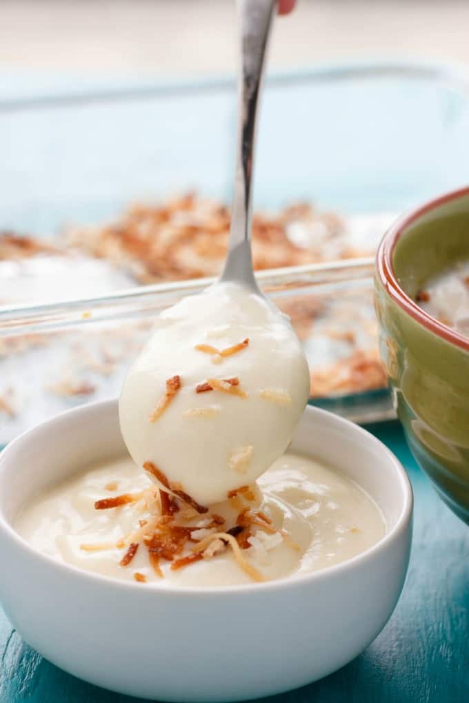 Homemade toasted coconut pudding in white bowl with spoon, green bowl and glass container on blue table