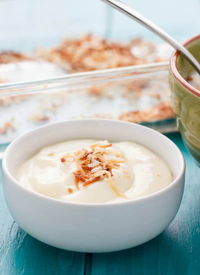 Homemade Toasted Coconut Pudding in white bowl on blue table next to green bowl and container in the background