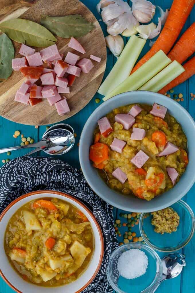 Slow Cooker Split Pea Soup with Ham in  bowls on blue table with vegetable, ingredients in small bowls, chopped ham on wooden pad and spoons