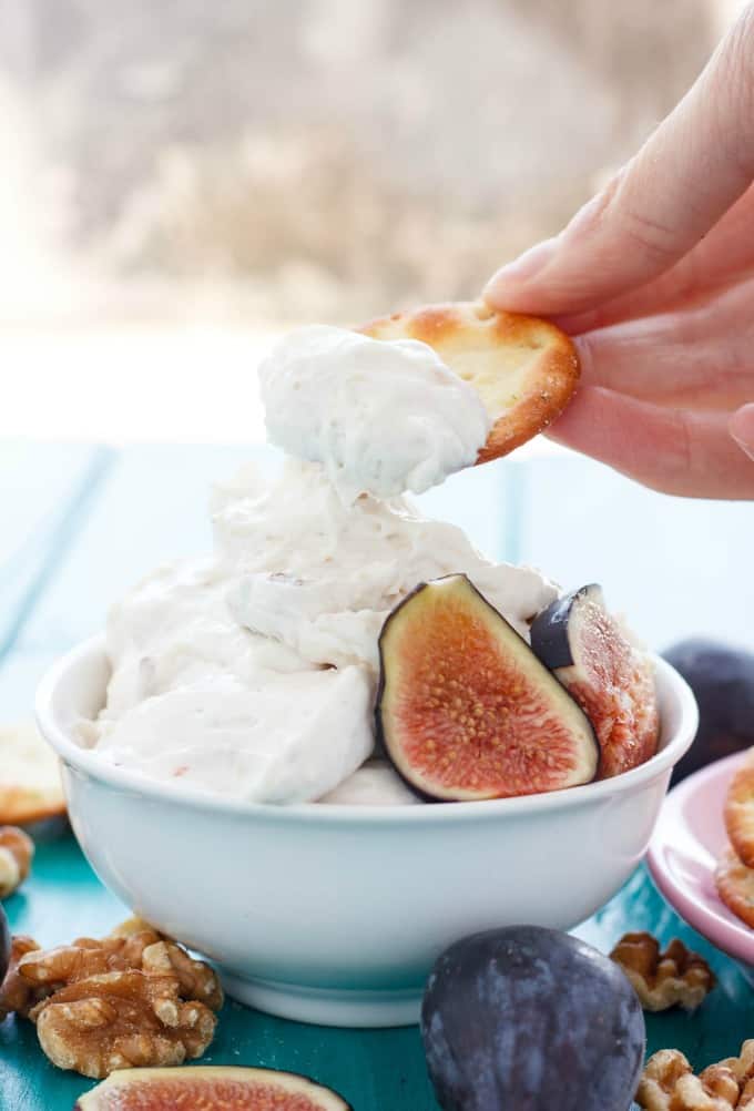 Farm Boy Fig and Walnut Cream Cheese Dip in white bowl picked by cracker held by hand on blue table with walnuts, figs and cracker