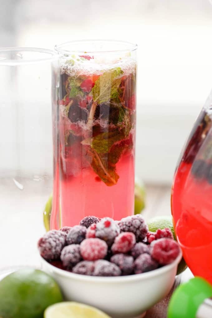 Mixed Berry Mojitos in glass cup and glass pitcher with frozen raspberries in bowl and limes on the table