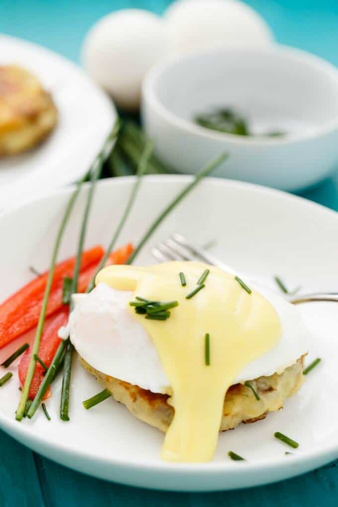 Eggs benedict over potato pancake on white plate with fork and vegetable