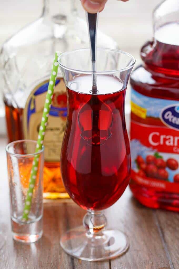 The Executioner drink in glass cup with spoon on table with glass shot with straw, cranberry juice in container and liquor in glass bottle