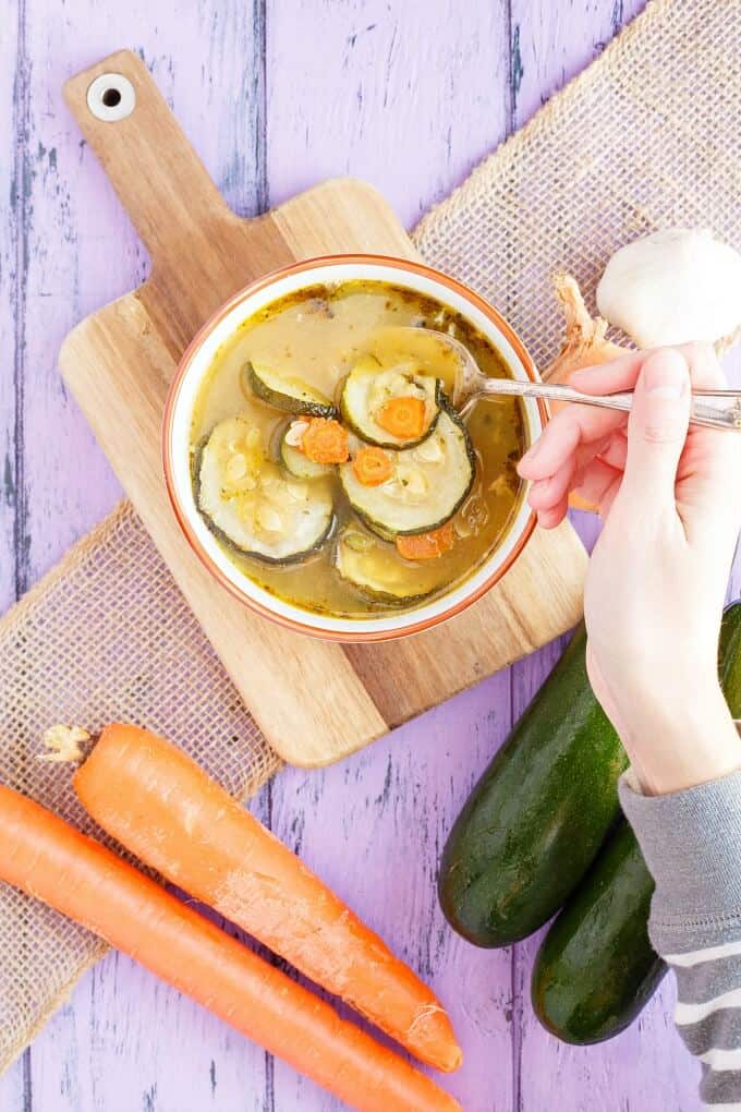 Roasted Zucchini-Carrot Soup in white bowl with soup held by hand on wooden pad on purple table with vegetables